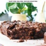 a healthy banana brownies with a piece broken showing the fudgy texture, placed on a wooden board and a green plant in the background with a glass of milk