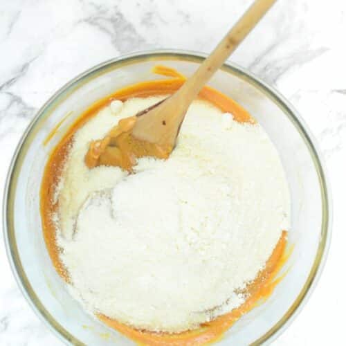 Coconut flour added to a bowl with a maple syrup and peanut butter mixture, with a wooden spoon.