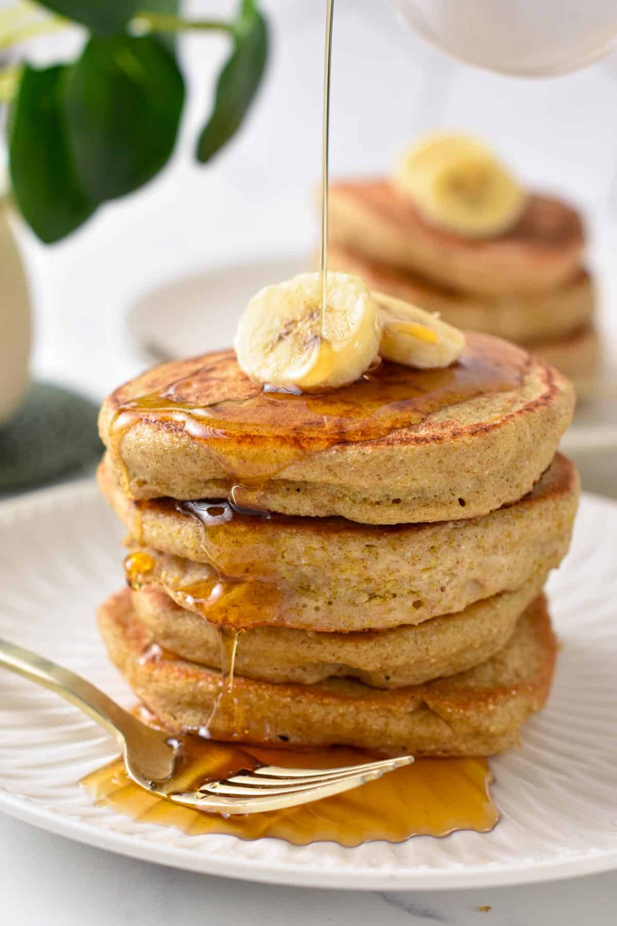 A stack of fluffy quinoa pancakes with banana slices on top and a drizzle of maple syrup.