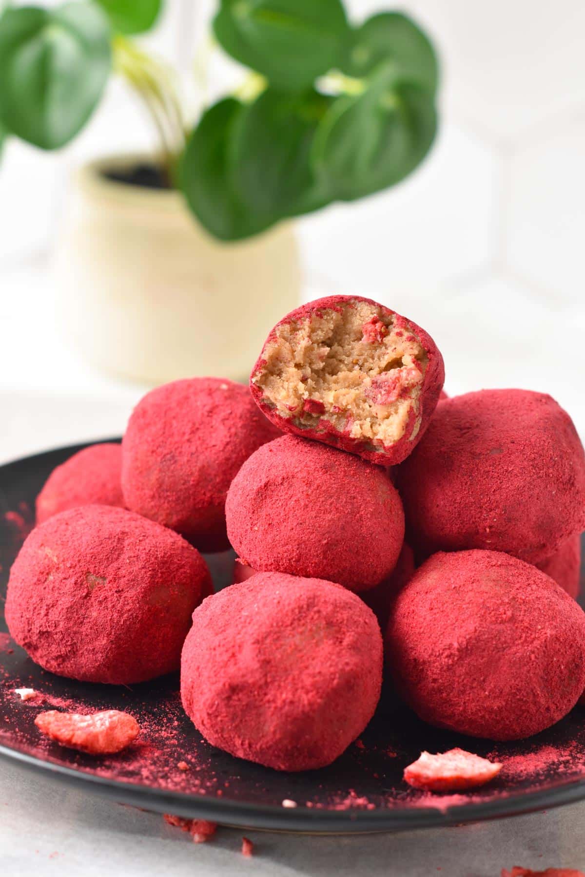 A black plate filled with a stack of pink Strawberry Protein Balls and the one on top half bitten showing the soft texture inside.