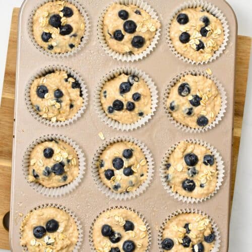 a 12 hole muffin pan filled with vegan oatmeal muffin batter and fresh blueberries on top