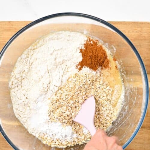 all the dry ingredients for a vegan oatmeal muffin recipe in a large glass bowl with a hand holding a pink spatula stirring the mixture