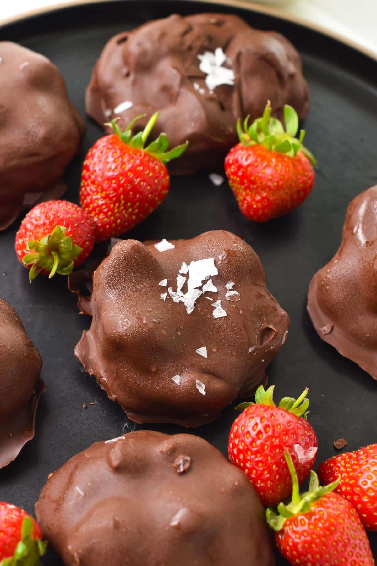 A black plate with chocolate coated strawberry yogurt clusters and fresh strawberries on the sides