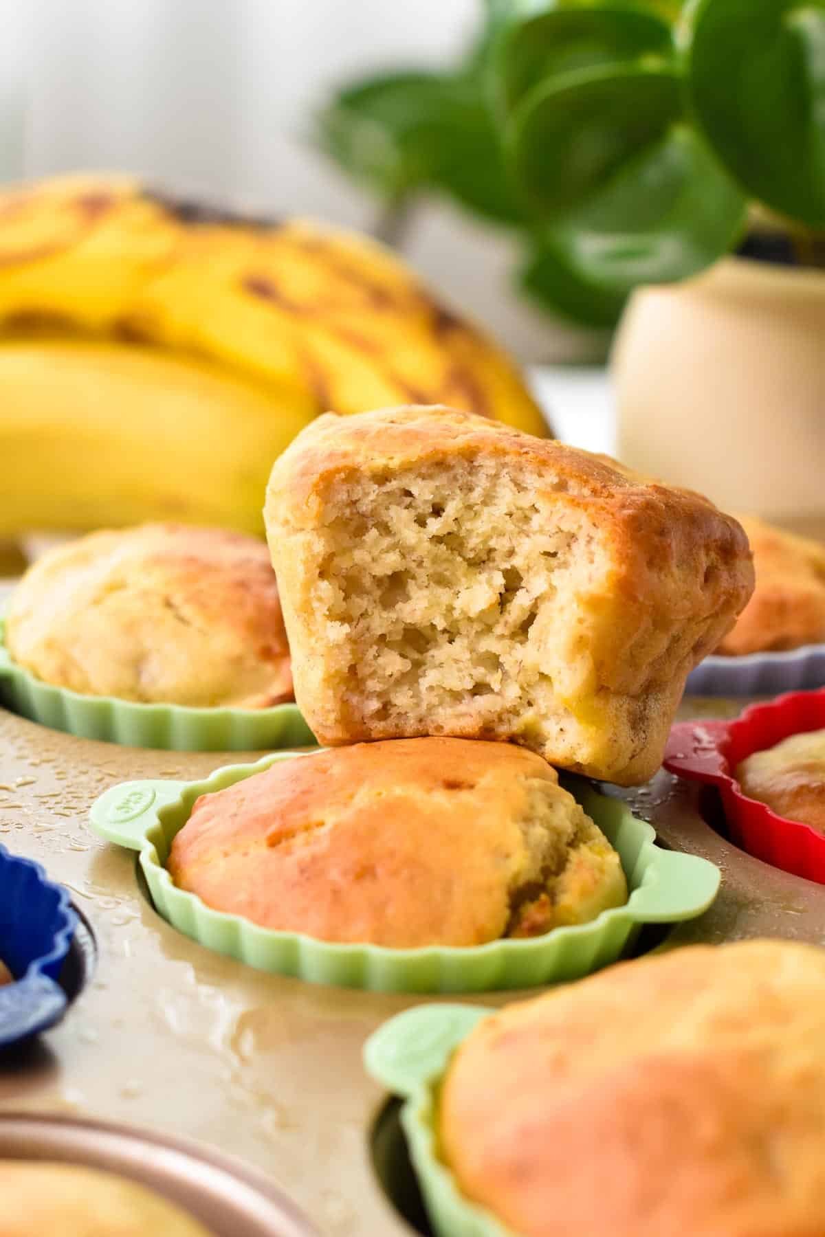 a banana muffins half beaten showing the fluffy texture inside and sitting on top of another banana muffins in its pale green silicone cup