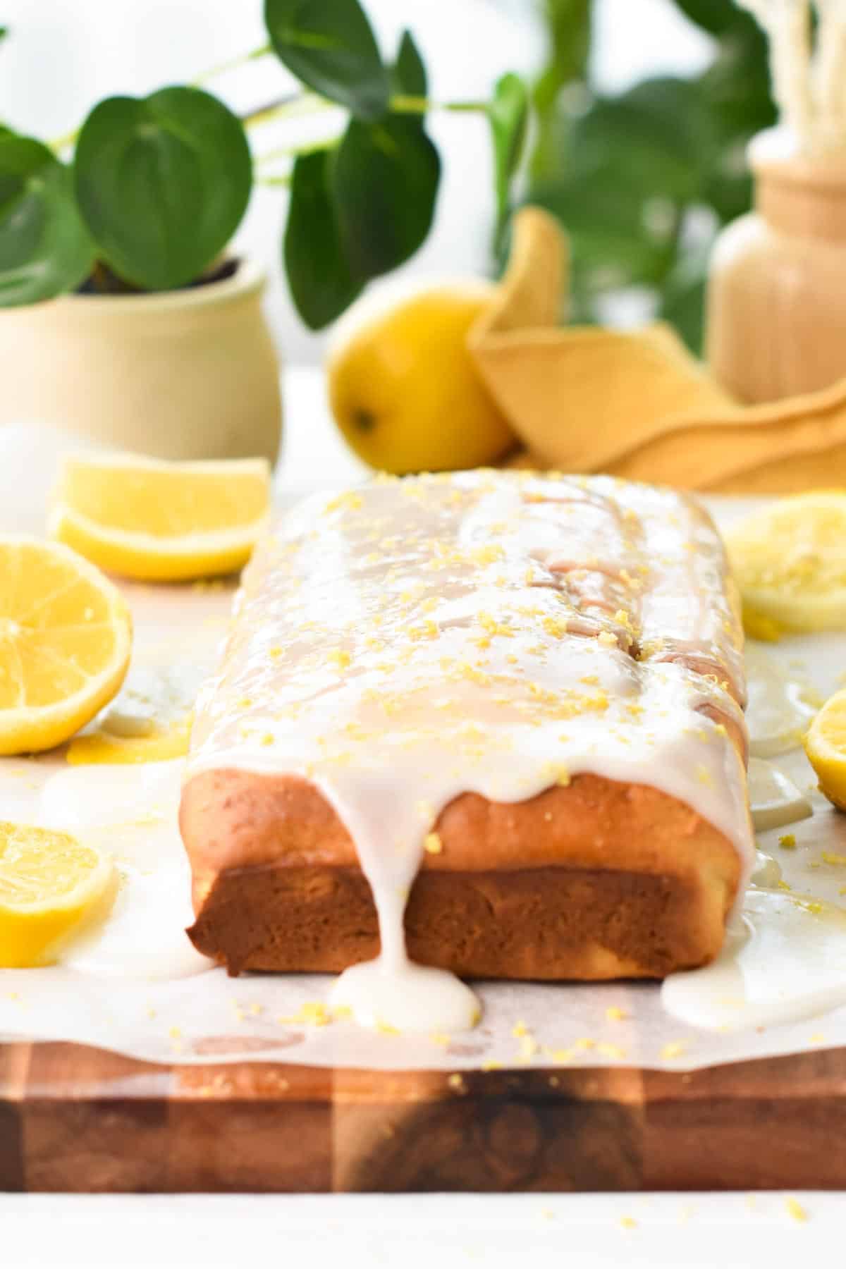 A glaze lemon pound cake on a chopping board with lemons and green plants in the background.