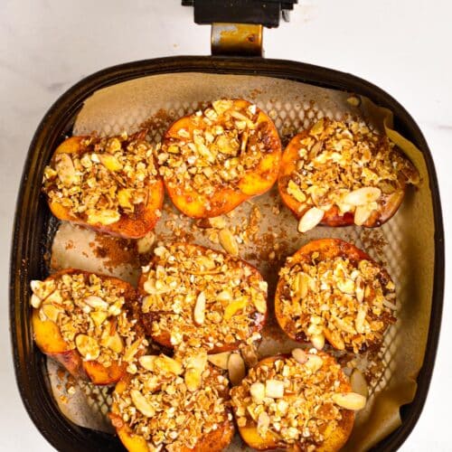 a air fryer basket lined with brown parchment paper and topped with halves peaches, covered with cinnamon sugar and oat crumble