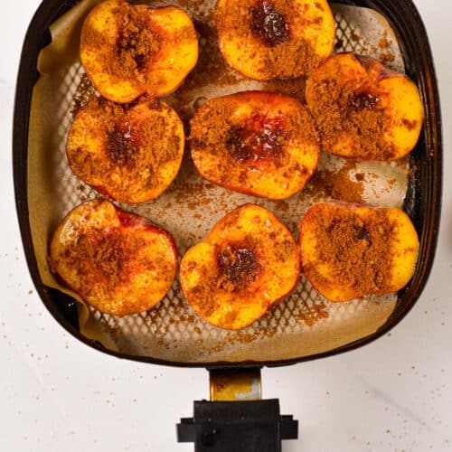 a air fryer basket lined with brown parchment paper and topped with halves peaches, covered with cinnamon sugar