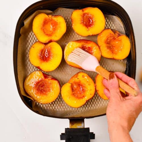 a air fryer basket lined with brown parchment paper and topped with halves peaches, with a hand brushing coconut oil on top of the peach flesh