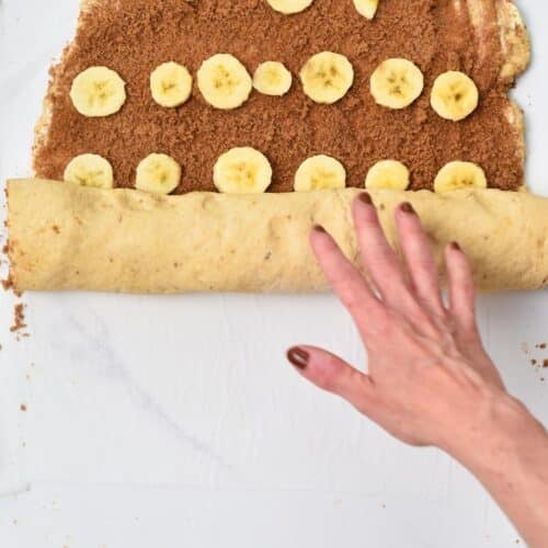 a hand rolling a rectangle cinnamon roll dough filled with cinnamon sugar and banana slices