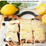 a blueberry lemon blondie with icing, cut into 9 squares on a wooden chopping board with lemons and blueberries around