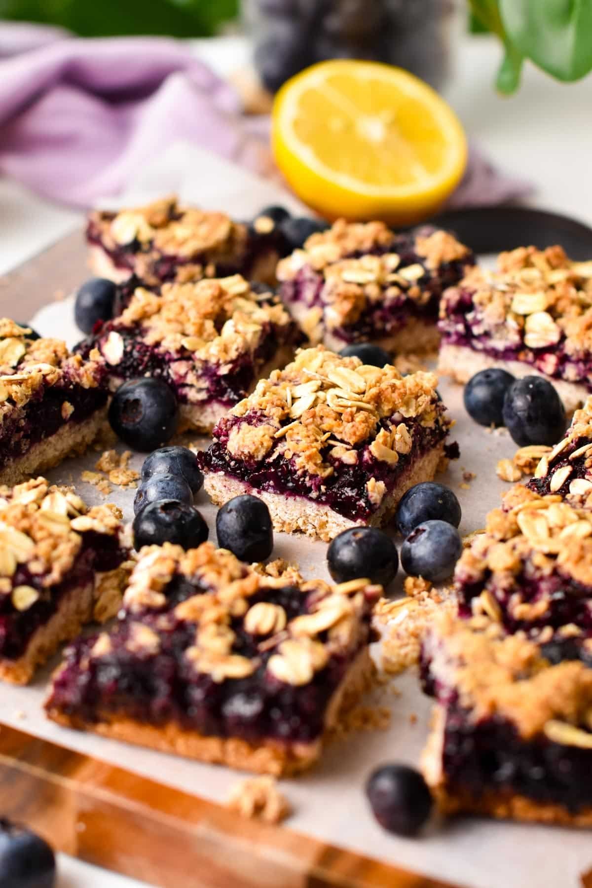 Squares of blueberry oatmeal bars on a chopping board with blueberries around and a lemon in the background.