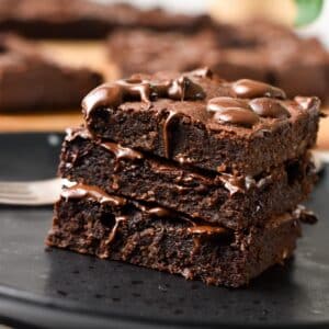 a stack of 3 slices of brownies on a black plate and melting chocolate chips on top