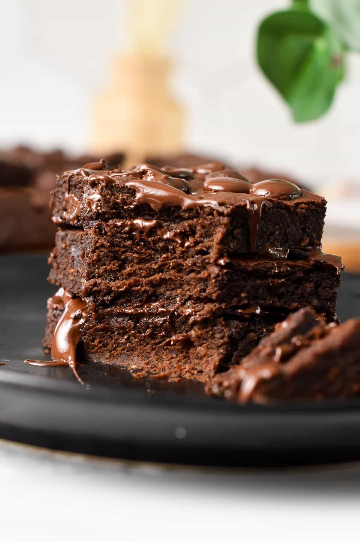 A stack of 3 slices of brownies on a black plate and melting chocolate chips on top.