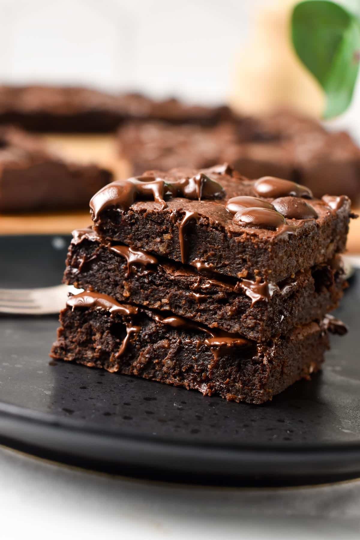 A stack of 3 slices of brownies on a black plate and melting chocolate chips on top.