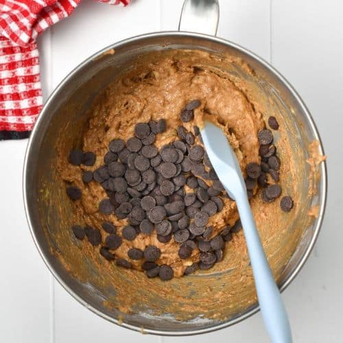 Chocolate chips on the Banana Almond Butter Muffin batter in a mixing bowl with a silicone spatula.