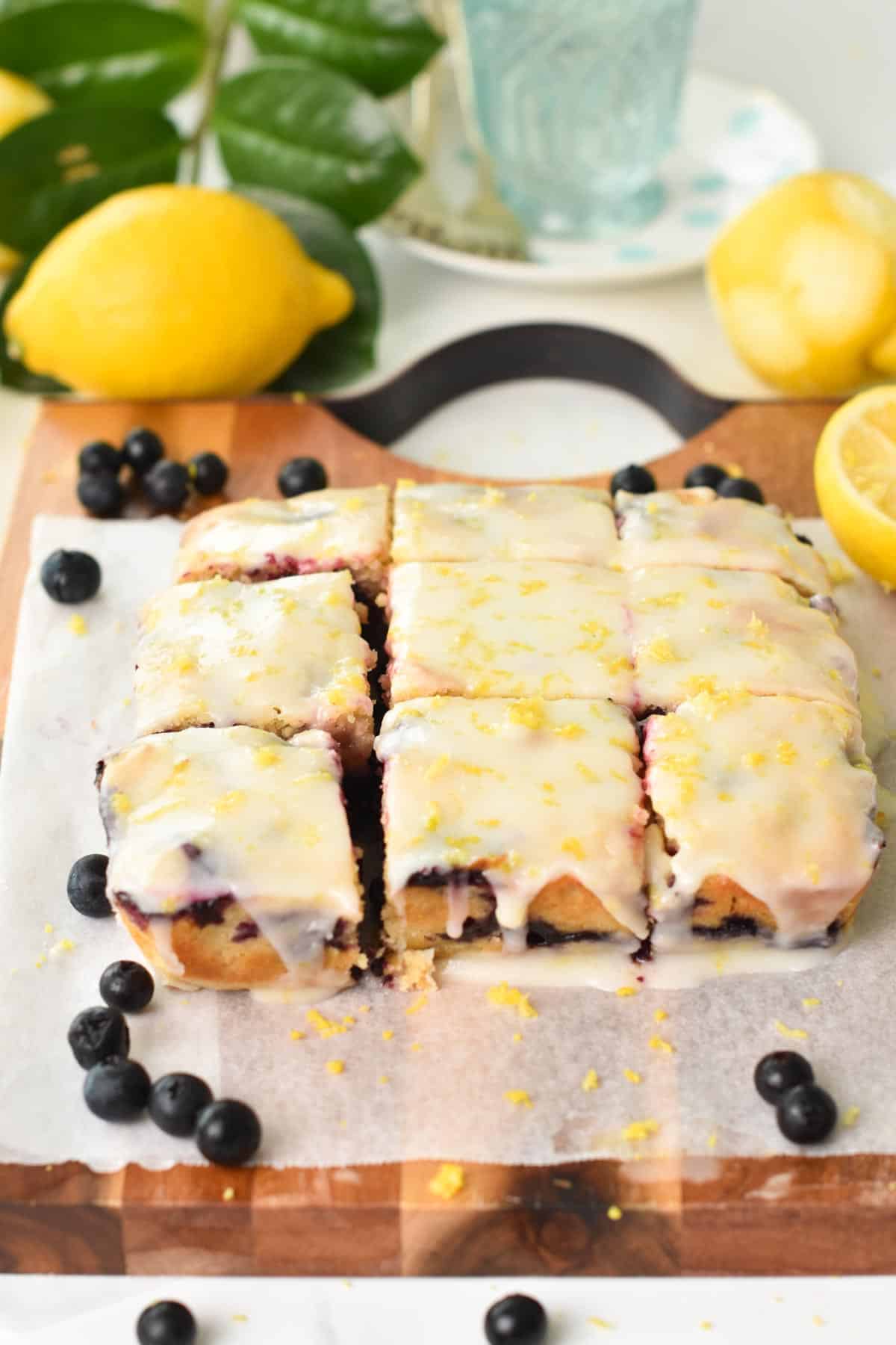 A blueberry lemon blondie with icing, cut into 9 squares on a wooden chopping board with lemons and blueberries around.