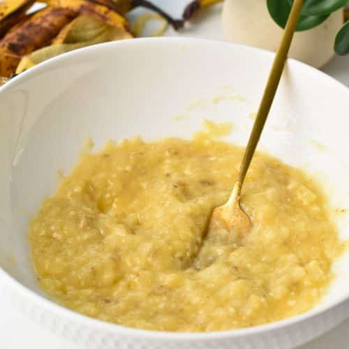 Mashed bananas in a mixing bowl with a golden spoon.