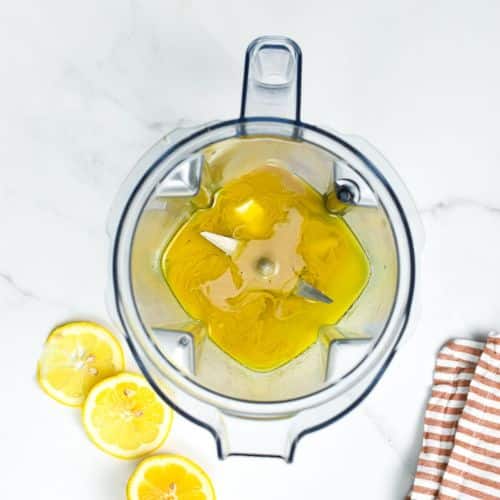 Tahini paste, olive oil, lemon juice, and garlic cloves in the jug of a blender.