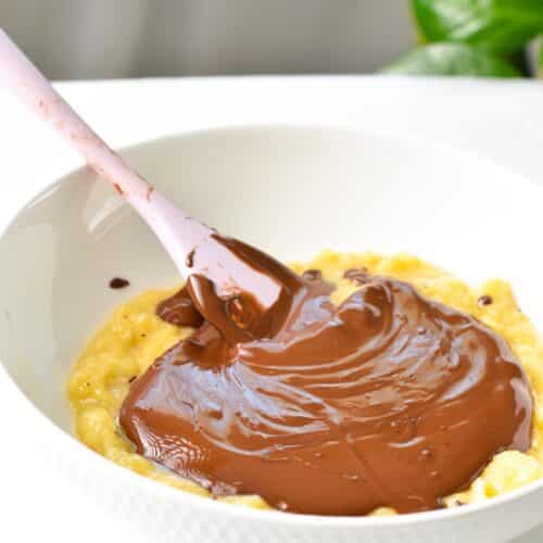 a mixing bowl filled with mashed banana and melted chocolate on top with a pink silicone spatula in the bowl