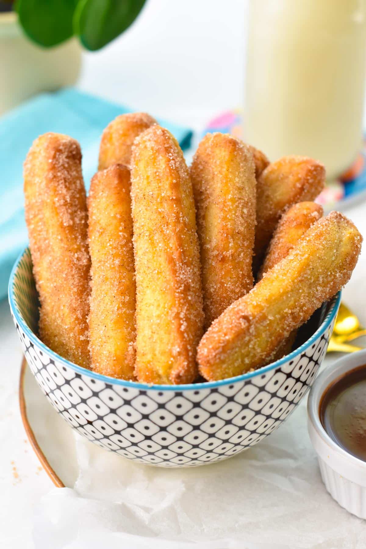 A bowl filled with golden air fryer churros coated with cinnamon sugar and a blue towel in the background.