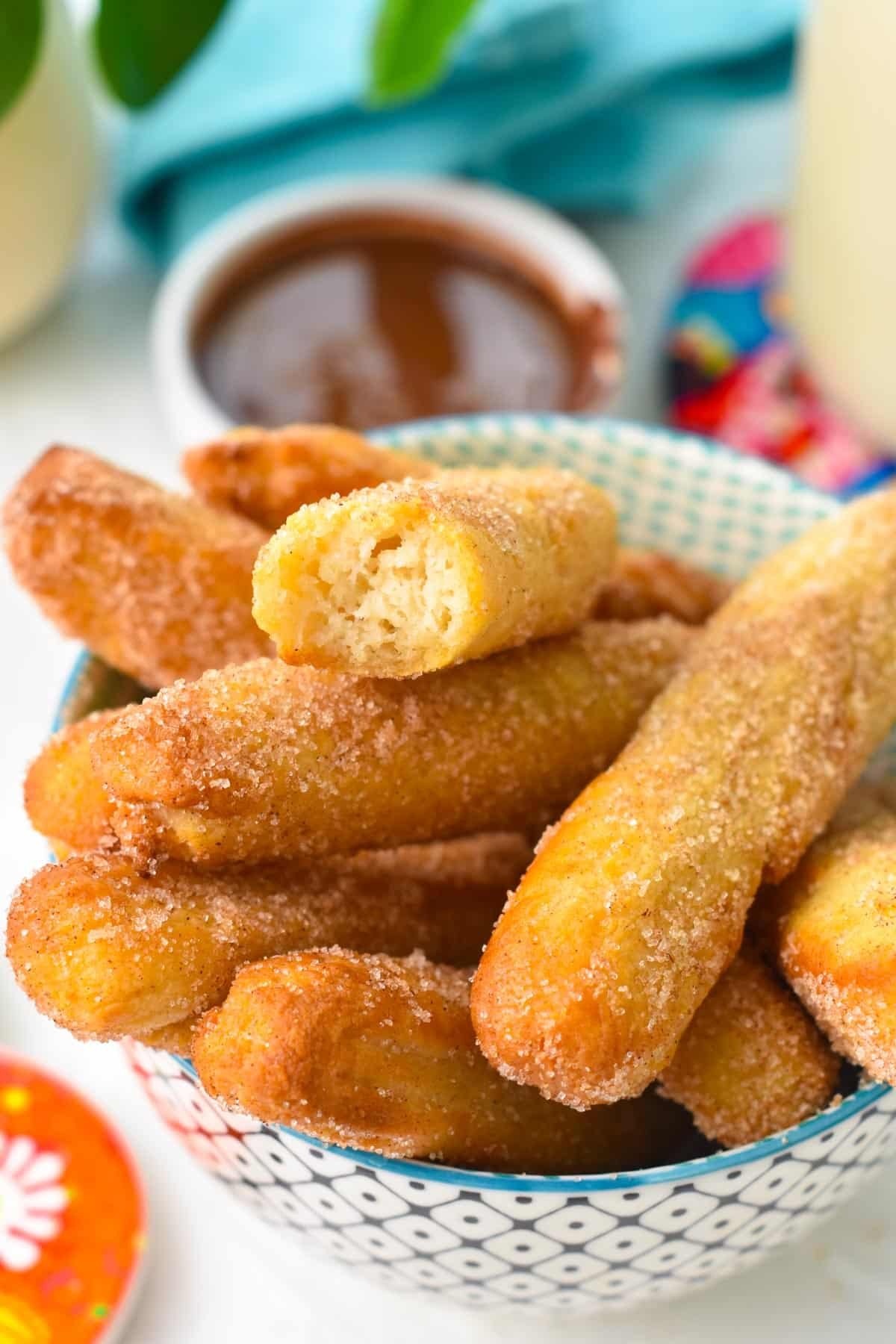 a bowl filled with golden air fryer churros coated with cinnamon sugar, one on top half eaten showing the airy texture of the churros, and chocolate sauce in the background