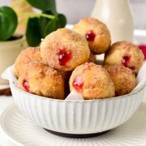 a bowl filled with jelly filled donut holes coated with cinnamon sugar and a green plant in the background