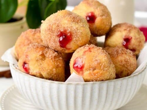 a bowl filled with jelly filled donut holes coated with cinnamon sugar and a green plant in the background
