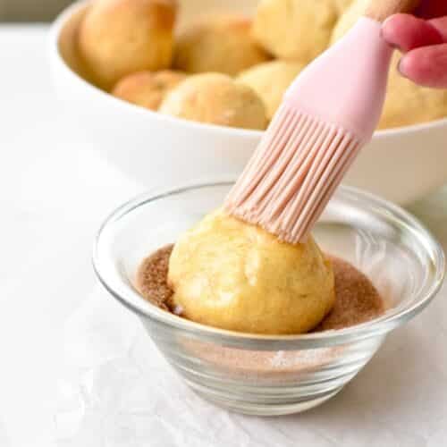 An air fried donut hole in a  bowl filled with cinnamon sugar and a pink rubber pastry brush brushing oil on top of the donut.