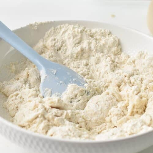 A bowl filled with a crumbly dough made of flour and yogurt, and a light blue rubber spatula in the bowl.