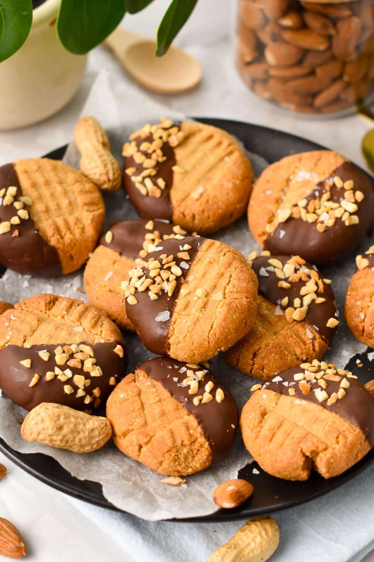 A black plate lined with white parchment paper and filled with lots of peanut butter cookies, half dipped in dark chocolate and covered with crushed peanuts.