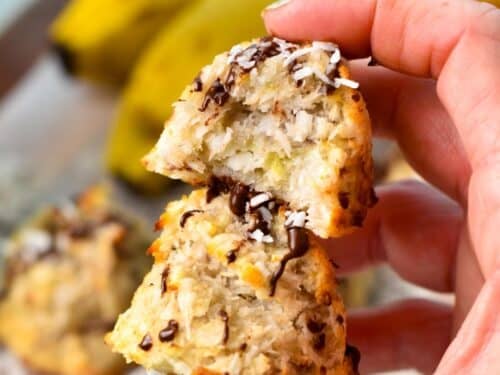 a hand holding two Coconut Banana Cookies with chocolate drizzle on to, stacked together with the one on top half eaten showing the texture inside
