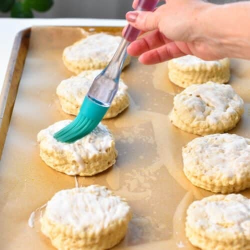 A baking sheet covered with parchment paper, and round cut scones on it plus a blue pastry brush brushing the top of the scones with yogurt.
