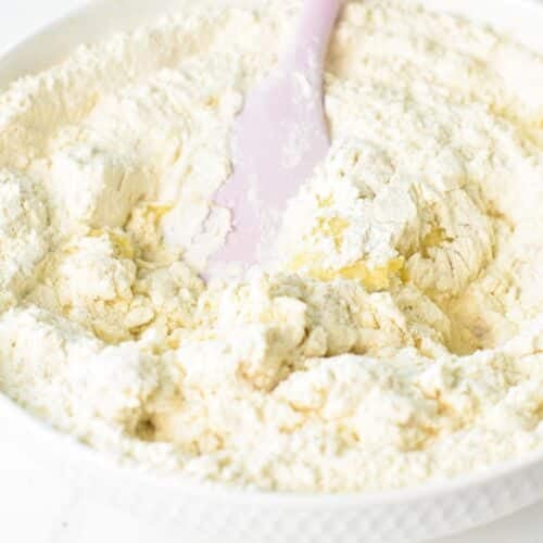 A mixing bowl with mashed bananas, yogurt, and flour and a pink spatula stirring the ingredients together.