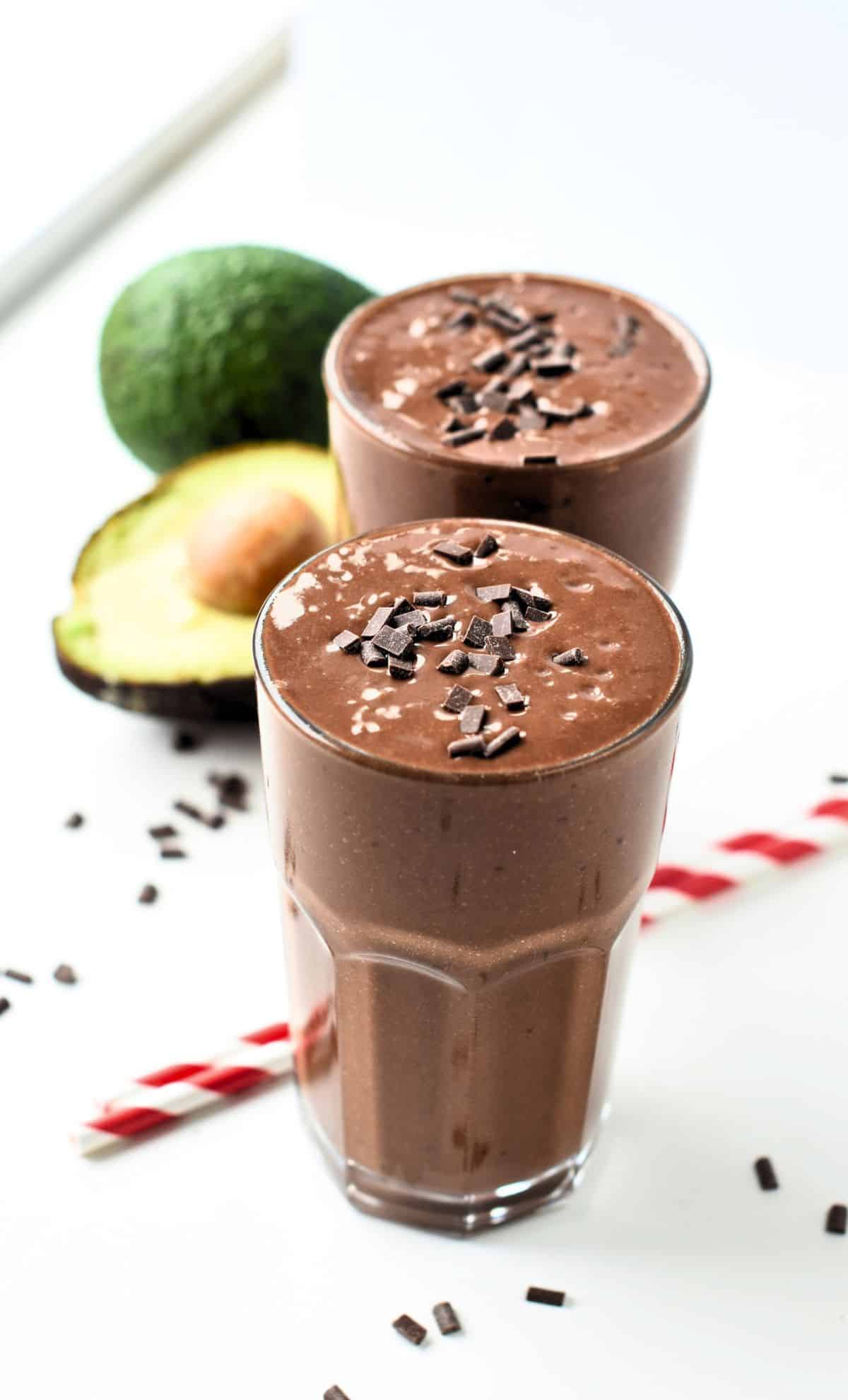 Two Chocolate Avocado Smoothie in tall glasses next to a sliced avocado.