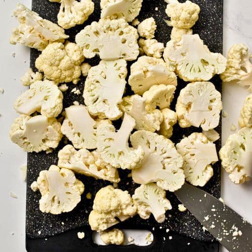Cut cauliflower florets on a chopping board with a kitchen knife.