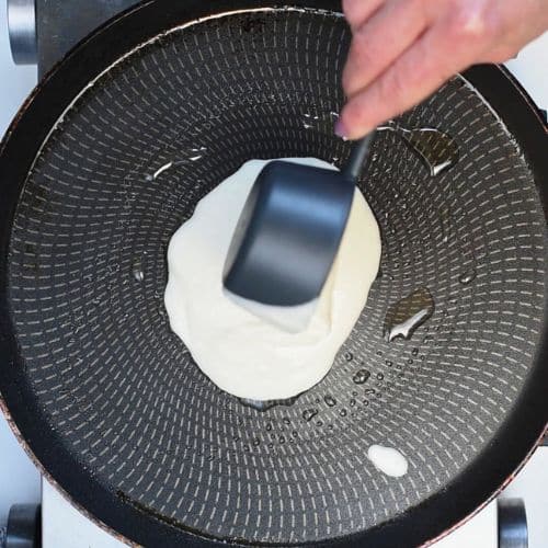 Pouring pancake batter from a cup onto a hot crepe pan.
