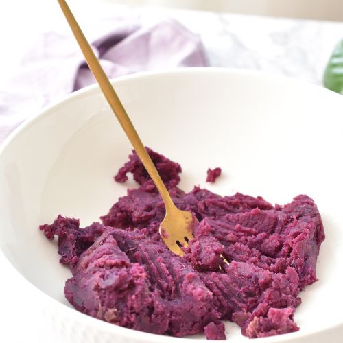 Boiled purple sweet potatoes turned into mash in a white bowl with a golden fork.